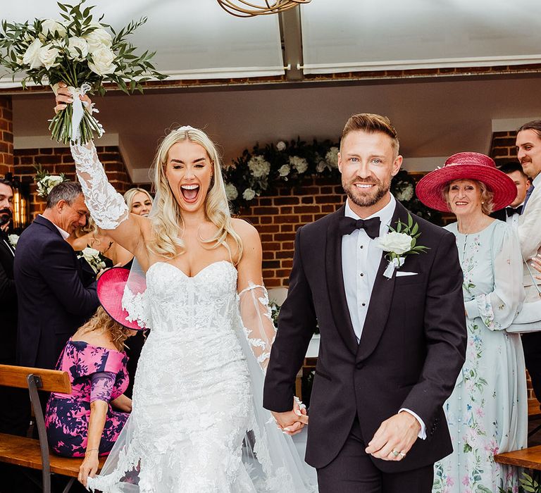 bride-in-lace-mermaid-wedding-dress-and-gloves-holding-white-rose-bouquet-and-groom-in-black-tuxedo