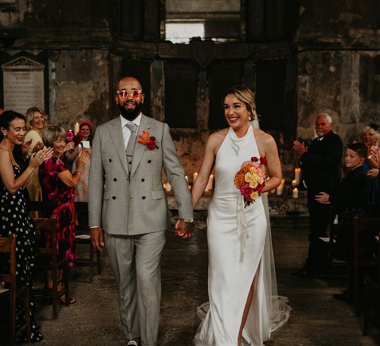 groom-in-grey-wedding-suit-with-bride-in-satin-wedding-dress-walking-back-down-the-aisle-as-married-couple