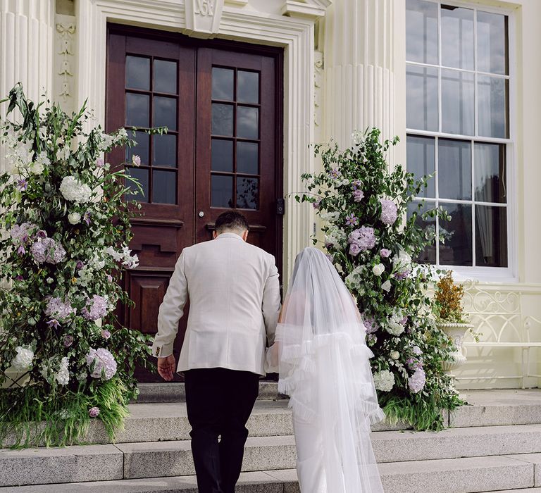 purple-wedding-flowers-at-country-house-with-bride-and-groom-walking-hand-in-hand