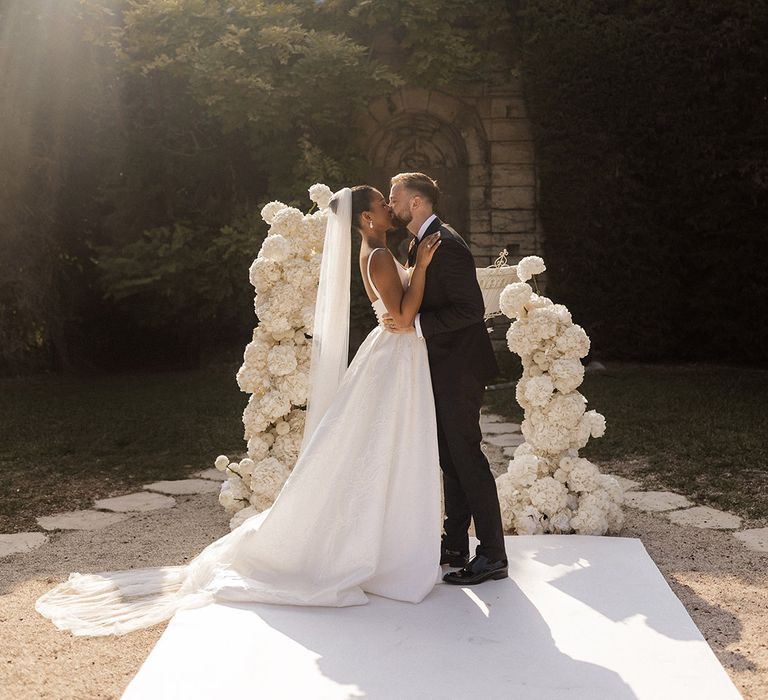 bride-and-groom-share-first-kiss-as-married-couple-in-front-of-white-rose-flower-columns