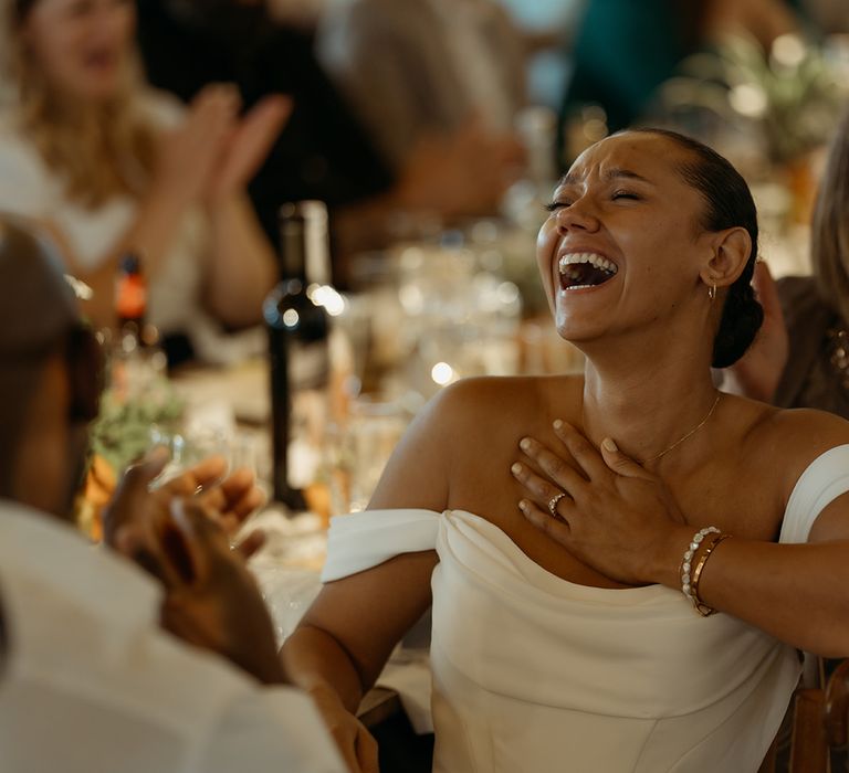 bride-laughing-in-off-the-shoulder-wedding-dress-listening-to-wedding-speeches