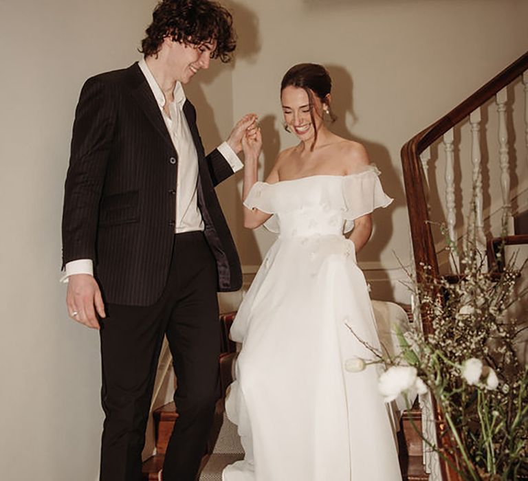 bride coming down the stairs with groom while wearing off-shoulder maxi wedding dress