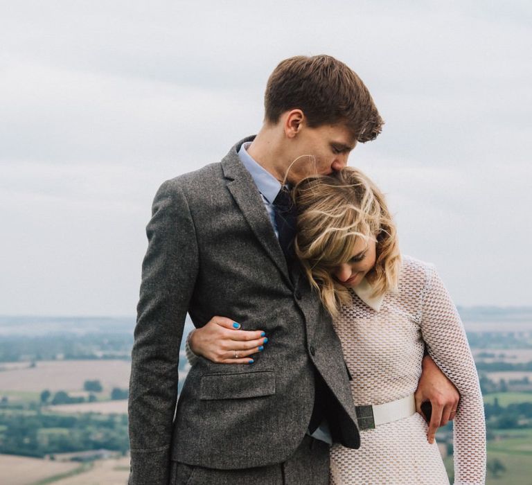 Bride and Groom stand side by side with arms around each other, groom kisses bride on the crown of her head