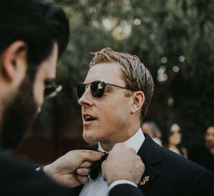 Groomsman with dark beard adjusts the bow tie on a white groom wearing sunglasses