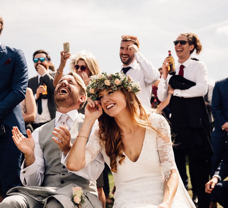 Groom in grey waistcoat laughs and claps hands with bride wearing flower crown next to him and guests smiling behind - Documentary Style Wedding Photography