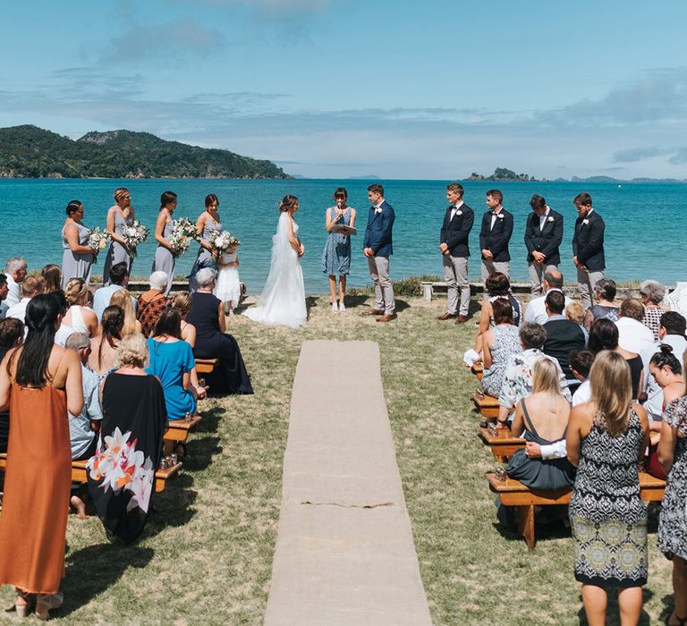 Wedding Ceremony | Bride in Estelle Sally Eagle Gown | Grooms in Grey Chinos & Navy Blazer | Outdoor Coastal Wedding at Ohawini Bay in New Zealand with Natural Garden Party Reception | Miss Gen Photography