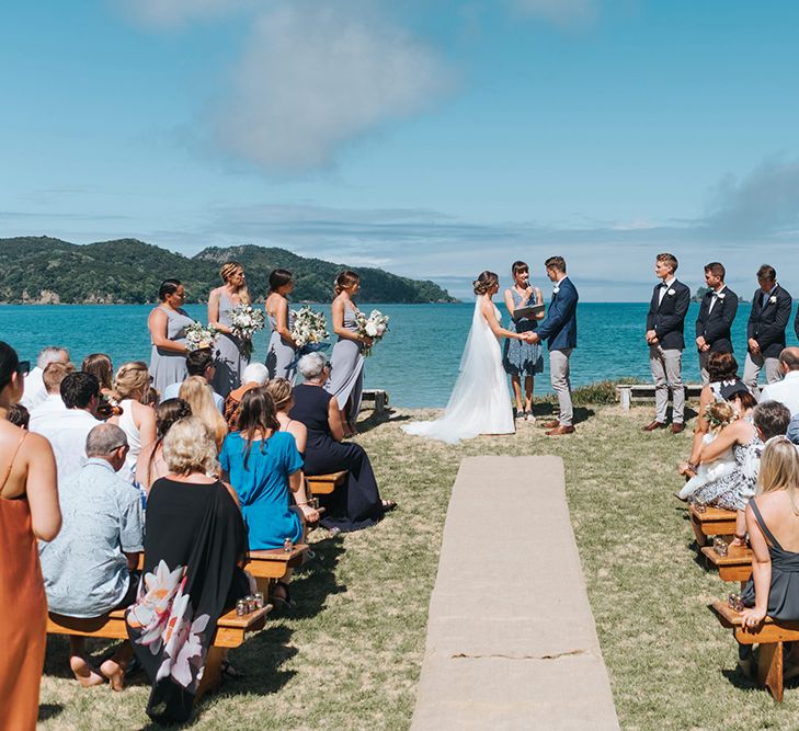 Wedding Ceremony | Bride in Estelle Sally Eagle Gown | Grooms in Grey Chinos & Navy Blazer | Outdoor Coastal Wedding at Ohawini Bay in New Zealand with Natural Garden Party Reception | Miss Gen Photography