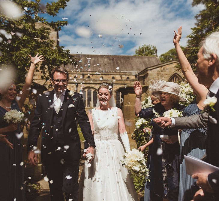 Bride in Tom Flowers Dress, Long Satin Gloves & Ivory & Co. Headdress and Groom in Moss Bros Suit at Church Confetti Exit