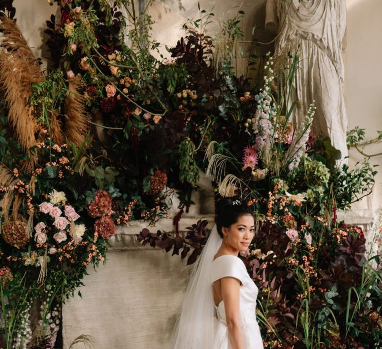 Bride in off the shoulder wedding dress standing in front of flower display at Aynhoe Park