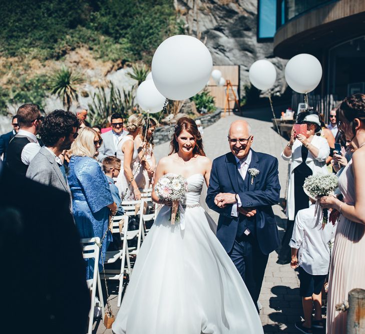 Bride in Strapless Ballgown Dress by Pronovias with Crystal Belt | Bridal Bouquet of Pink Gerberas, White Gerberas and Gypsophila Tied with Pink Trailing Ribbon | Father of the Bride in Navy Three Piece Suit with Pink Tie | Oversized White Balloons with Foliage String | Arrival of the Bride | Gypsophila Arch and Giant Balloons for an Outdoor Coastal Wedding | Toby Lowe Photography