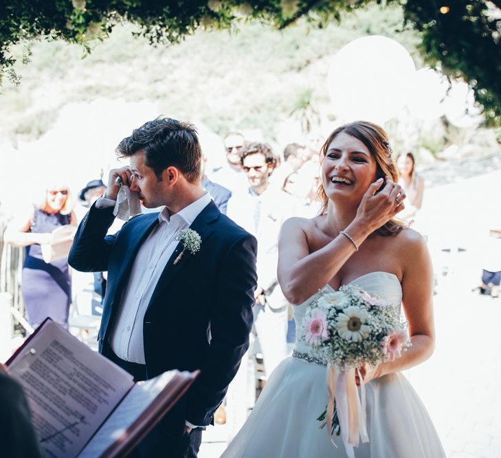 Bride in Strapless Ballgown Dress by Pronovias with Crystal Belt | Bridal Bouquet of Pink Gerberas, White Gerberas and Gypsophila Tied with Pink Trailing Ribbon | Groom in Navy Suit with Gypsophila Buttonhole | Gypsophila Wedding Arch with Fairy Lights | Wedding Ceremony Emotions | Gypsophila Arch and Giant Balloons for an Outdoor Coastal Wedding | Toby Lowe Photography