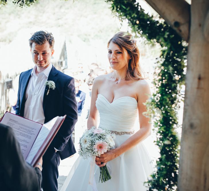 Bride in Strapless Ballgown Dress by Pronovias with Crystal Belt | Bridal Bouquet of Pink Gerberas, White Gerberas and Gypsophila Tied with Pink Trailing Ribbon | Groom in Navy Suit with Gypsophila Buttonhole | Gypsophila Wedding Arch with Fairy Lights | Wedding Ceremony | Gypsophila Arch and Giant Balloons for an Outdoor Coastal Wedding | Toby Lowe Photography