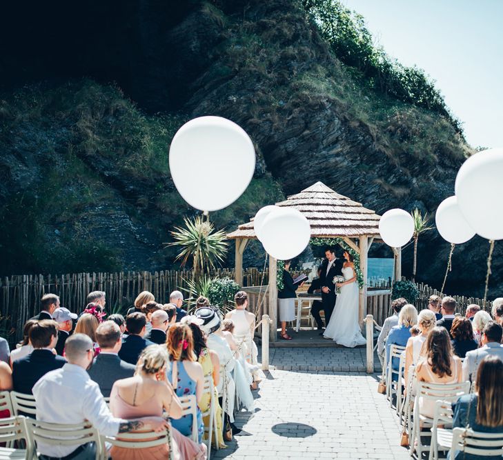 Oversized White Balloons with Foliage String | Bride in Strapless Ballgown Dress by Pronovias with Crystal Belt | Bridal Bouquet of Pink Gerberas, White Gerberas and Gypsophila Tied with Pink Trailing Ribbon | Groom in Navy Suit with Gypsophila Buttonhole | Gypsophila Wedding Arch with Fairy Lights | Wedding Ceremony | Gypsophila Arch and Giant Balloons for an Outdoor Coastal Wedding | Toby Lowe Photography