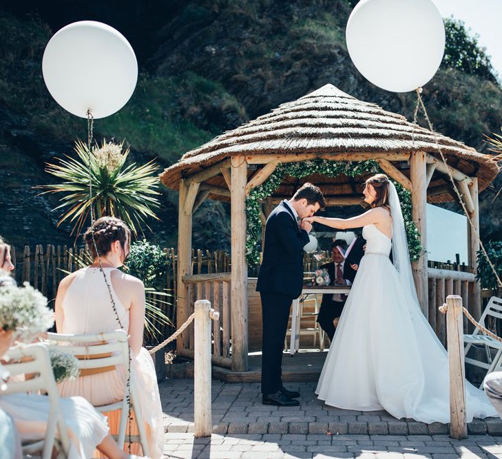 Bride in Strapless Ballgown Dress by Pronovias with Crystal Belt | Fingertip Veil | Groom in Navy Suit with Gypsophila Buttonhole | Oversized White Balloons with Foliage String | Gypsophila Wedding Arch with Fairy Lights | Wedding Ceremony | Gypsophila Arch and Giant Balloons for an Outdoor Coastal Wedding | Toby Lowe Photography