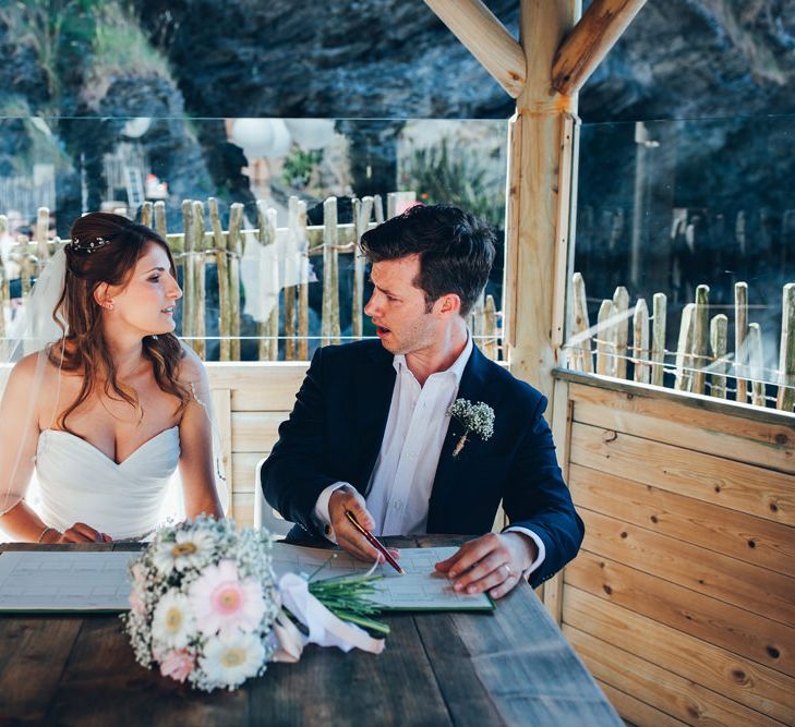 Bride in Strapless Ballgown Dress by Pronovias with Crystal Belt | Fingertip Veil | Bridal Bouquet of Pink Gerberas, White Gerberas and Gypsophila Tied with Pink Trailing Ribbon | Groom in Navy Suit with Gypsophila Buttonhole | Signing of the Register | Gypsophila Arch and Giant Balloons for an Outdoor Coastal Wedding | Toby Lowe Photography