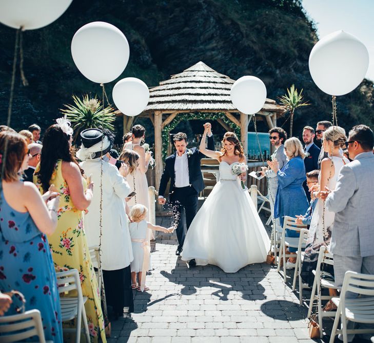 Bride in Strapless Ballgown Dress by Pronovias with Crystal Belt | Bridal Bouquet of Pink Gerberas, White Gerberas and Gypsophila Tied with Pink Trailing Ribbon | Groom in Navy Suit with Gypsophila Buttonhole | Oversized White Balloons with Foliage String | Gypsophila Wedding Arch | Just Married | Gypsophila Arch and Giant Balloons for an Outdoor Coastal Wedding | Toby Lowe Photography