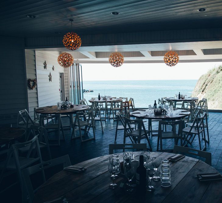 Wedding Reception by the Sea at Tunnels Beaches in Devon | Copper Lights | Round Wooden Tables | Gypsophila Arch and Giant Balloons for an Outdoor Coastal Wedding | Toby Lowe Photography