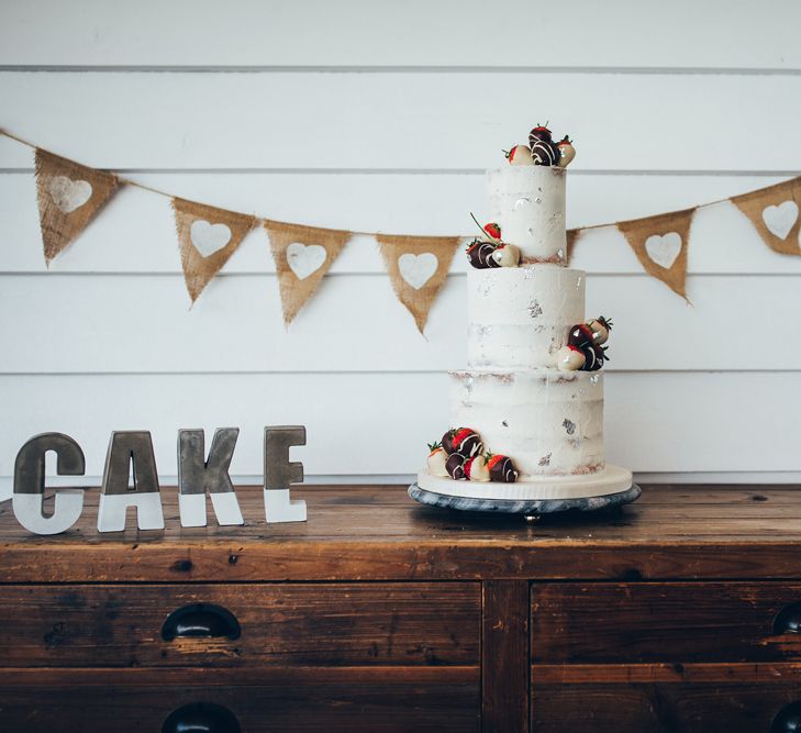 Three-Tier Semi-Naked Cake with Chocolate Dipped Strawberries | Hessian Bunting with White Hearts | Gypsophila Arch and Giant Balloons for an Outdoor Coastal Wedding | Toby Lowe Photography