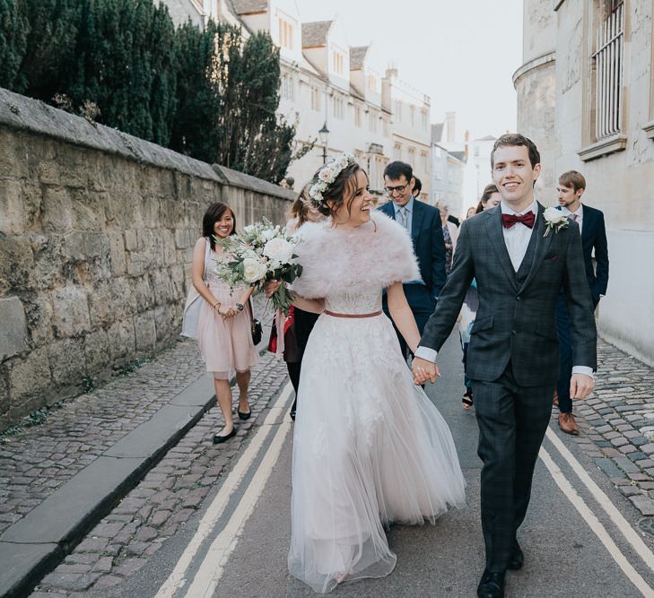 Bride and groom take a walk with their guests at an intimate Oxford wedding with a bridal shrug and a flower head crown