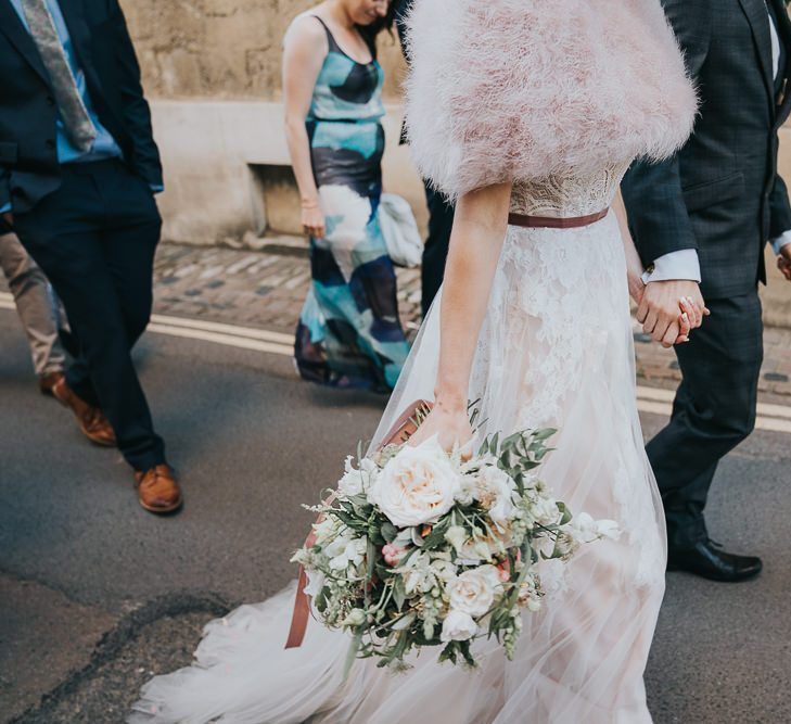 Bride and groom take a walk with their guests at an intimate celebration with a bridal shrug and a beautiful white floral bouquet