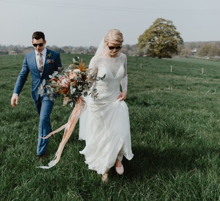 Bride in Lace Allure Bridals Wedding Dress with Long Sleeves and Groom in Blue Hugo Boss Suit Walking Through a Field