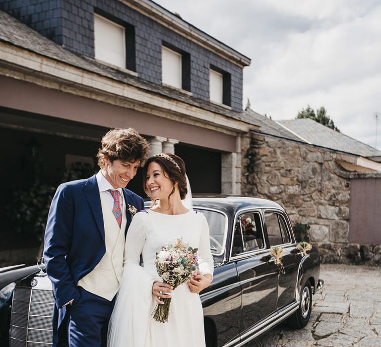 Bride in Nnavascues Embroidered Back Wedding Dress and Groom in Navy Suit Standing By Their Wedding Car