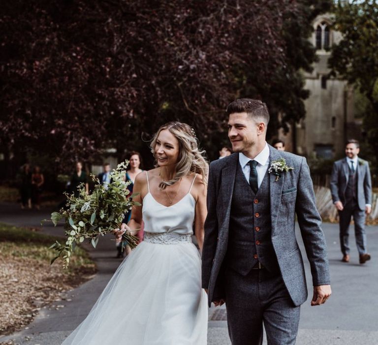 Bride and groom walk together to their pub reception wearing two piece gown and foliage bouquet