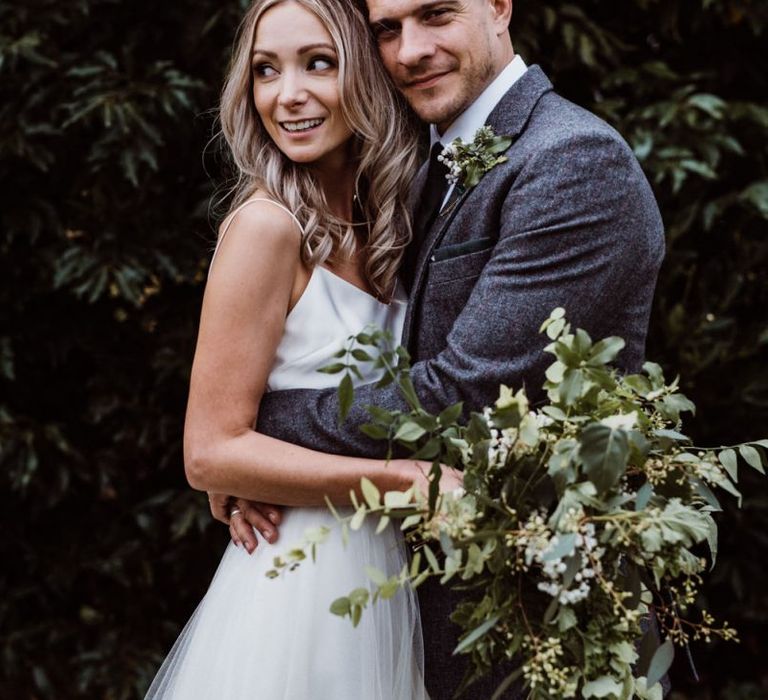 Bride and groom embrace whilst clutching foliage bouquet