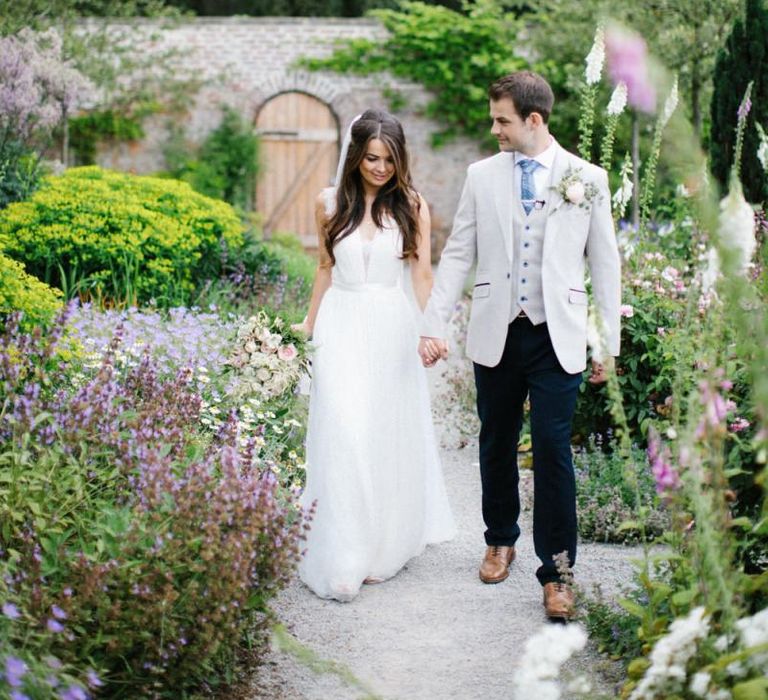 Bride in Catherine Deane Wedding Dress and Groom in Beige Blazer Holding Hands in Middle Lodge Estate's Gardens