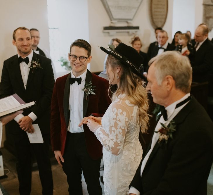 Groom in Red Velvet Jacket with Bow Tie and Glasses Smiling at His Bride During the Wedding Ceremony