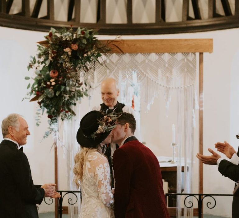 Bride in Phase Eight Wedding Dress and Fedora Hat and Groom in Red Velvet Jacket Kissing at the Altar