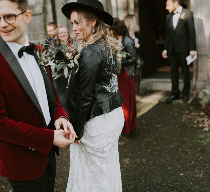 Bride in Fedora Hat and Personalised Leather Jacket  and Groom in Red Velvet Jacket Outside the Church