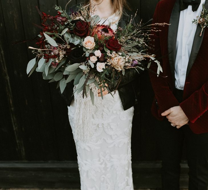 Bride in Phase Eight Wedding Dress Holding an Oversized Bouquet with Red Roses and Proteas and Groom in Red Velvet Tuxedo Jacket