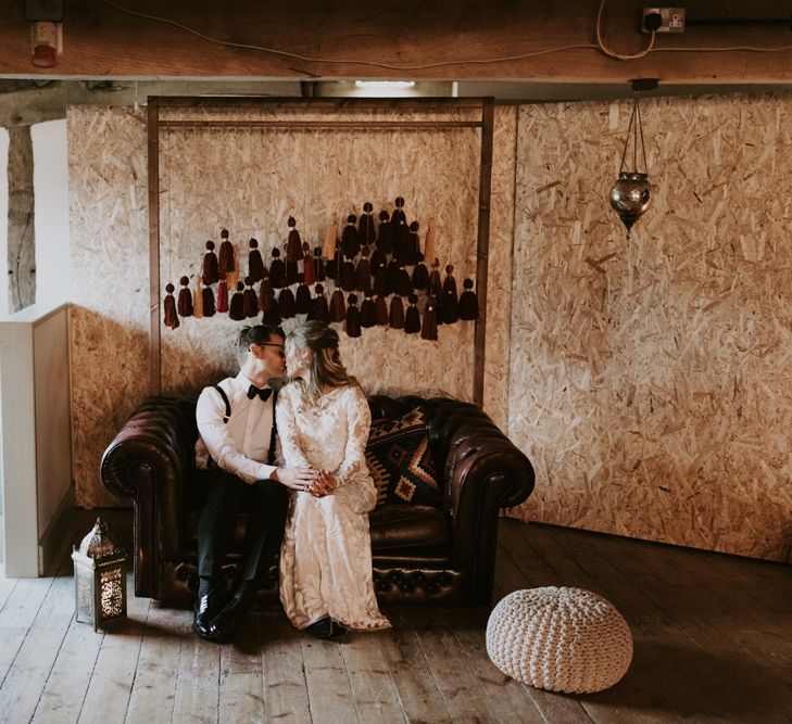 Bride and groom Sitting on a Leather rSofa with Wooden Frame Backdrop Decorated with Hanging Tassels