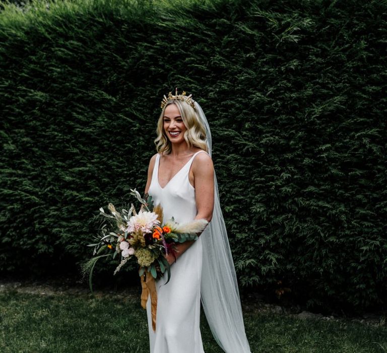 Stylish bride in slip wedding dress and gold bridal crown holding  an oversized bouquet