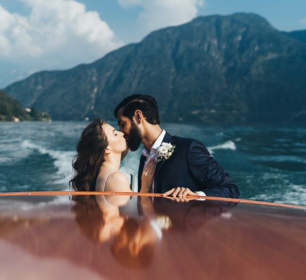 Bride in Grace Loves Lace Mia Wedding Dress and Groom in Taliare Navy Suit Kissing on Boat Ride Over Lake Como