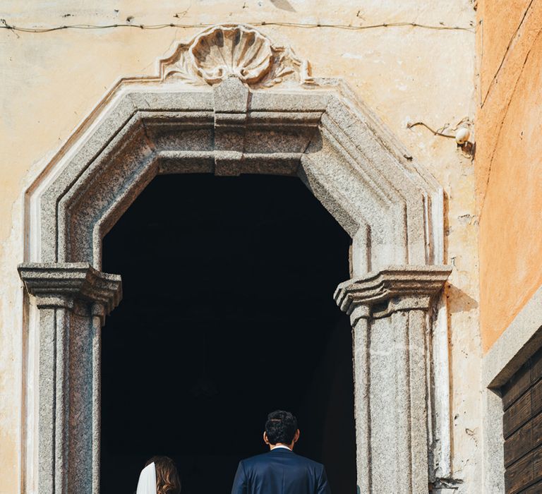 Bride in Grace Loves Lace Mia Wedding Dress and Groom in Taliare Navy Suit Entering Wedding Ceremony Venue