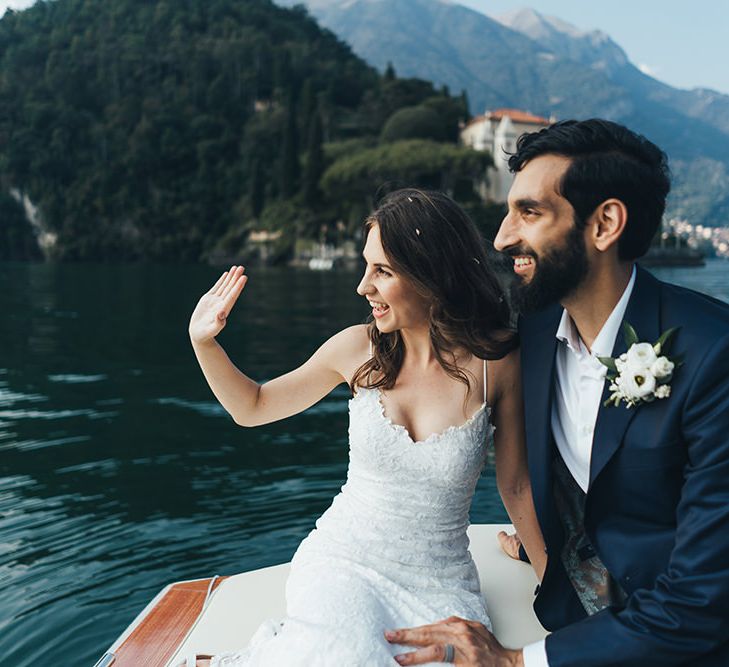 Waving Bride in Grace Loves Lace Mia Wedding Dress and Groom in Taliare Navy Suit Enjoying a Boat Ride Over Lake Como