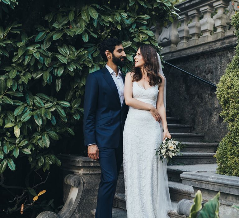 Bride in Grace Loves Lace Mia Wedding Dress and Groom in Taliare Navy Suit Embracing on Steps