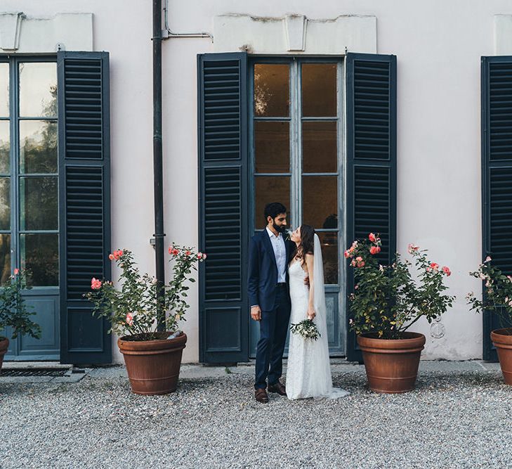 Bride in Grace Loves Lace Mia Wedding Dress and Groom in Taliare Navy Suit Standing in Front of giant Window Shutters