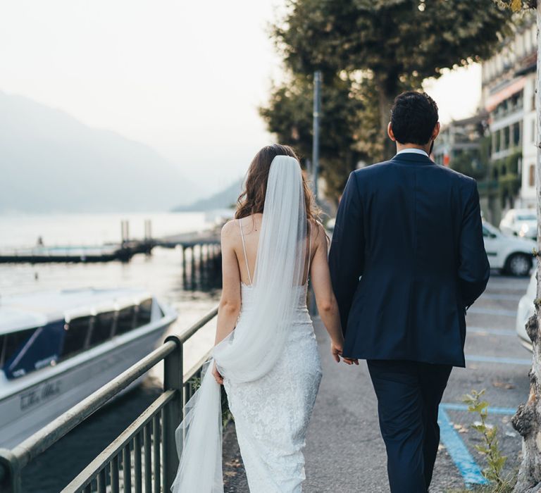 Bride in Grace Loves Lace Mia Wedding Dress and Groom in Taliare Navy Suit Walking Around Lake Como