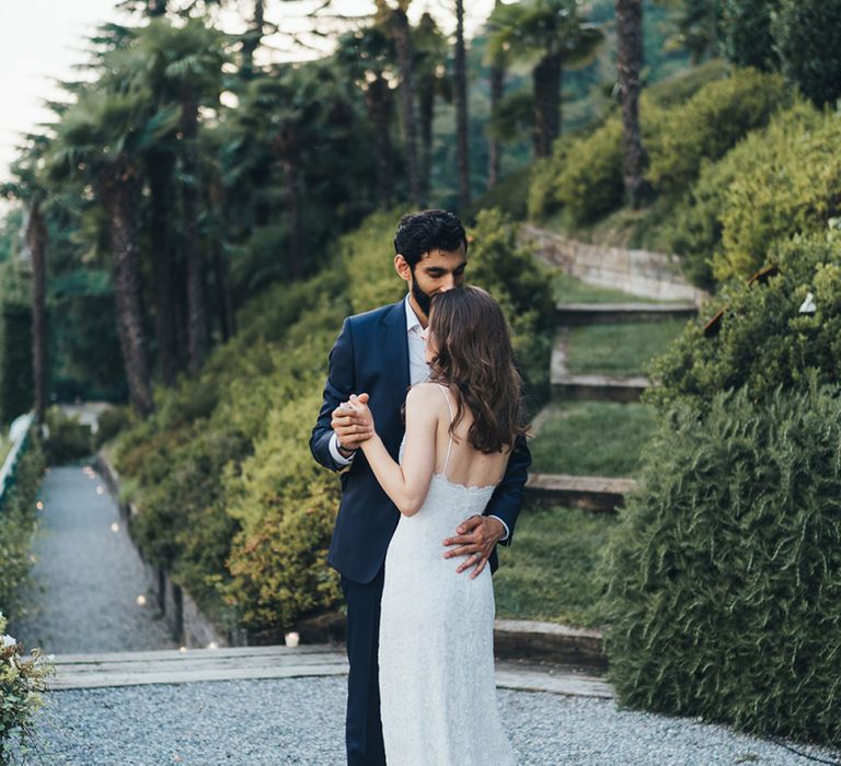 Bride in Grace Loves Lace Mia Wedding Dress and Groom in Taliare Navy Suit Embracing in Gardens