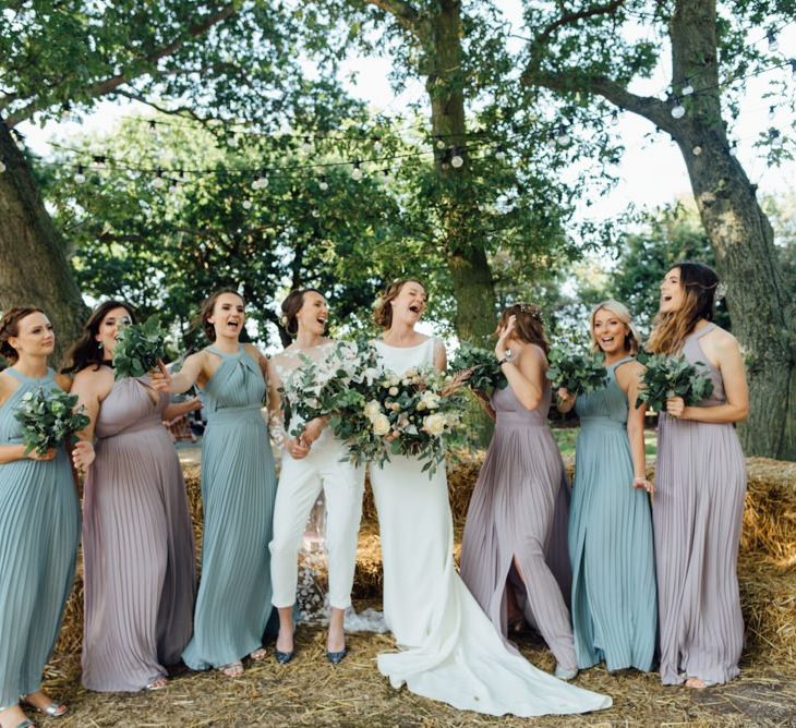 Bride in appliqué wedding jumpsuit and bride in elegant embellished gown with bridesmaids in pastel halter neck dresses at woodland celebration