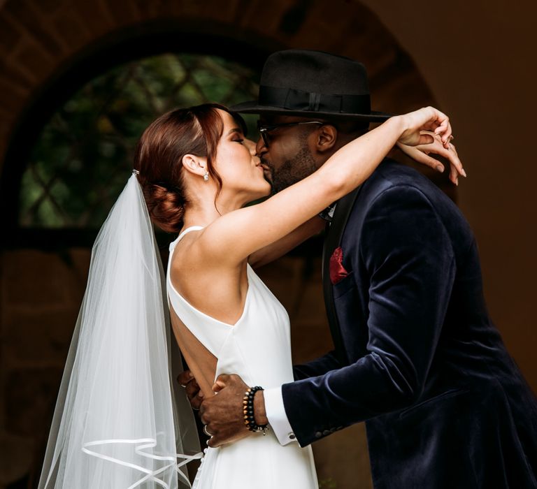 Groom in velvet dinner jacket and fedora kissing his bride at The Ravenswood wedding