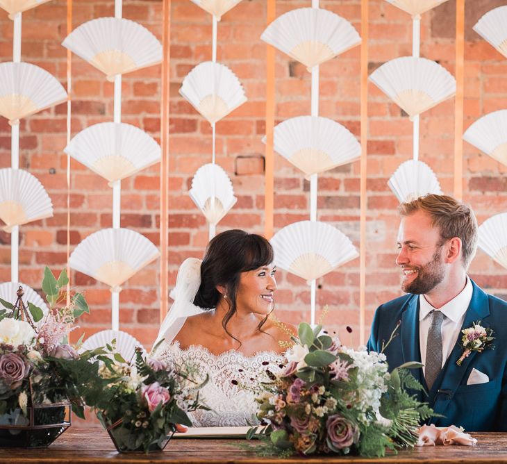 Signing of the Register with Backdrop of Chinese Paper Fans | Bride in Lace Watters Dress with Bardot Neckline and Buttons Down Back | Blossom and Bluebird Cathedral Length Veil | Bridal Up Do | Groom in Royal Blue Three-Piece Suit from Moss Bros. with Silver Tie and Pocket Square from T.M.Lewin | Bridal Bouquet of Dusky Pink Roses, White Peonies, Sweetpeas, Coffee-Coloured Phlox and Foliage Tied with Cappuccino Silk Ribbon | Wedding Ceremony at The West Mill | Smoke Bombs and Chinese Paper Fans Backdrop with Bride in Bardot Dress | Twig &amp; Vine Photography