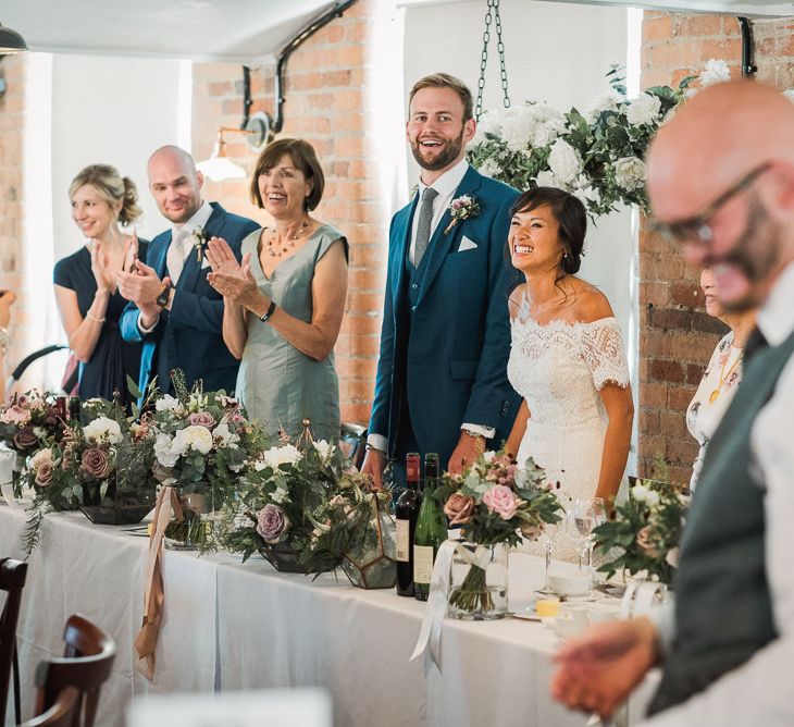 Bride in Lace Watters Dress with Bardot Neckline and Buttons Down Back | Bridal Up Do | Groom in Royal Blue Three-Piece Suit from Moss Bros. with Silver Tie and Pocket Square from T.M.Lewin | Hanging Floral Installation Over Top Table | Dusky Pink Roses, White Peonies, Sweetpeas, Coffee-Coloured Phlox and Foliage in Hurricane Jars on Top Table | Wedding Reception at The West Mill |  Smoke Bombs and Chinese Paper Fans Backdrop with Bride in Bardot Dress | Twig &amp; Vine Photography