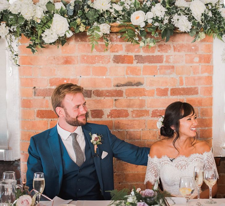 Bride in Lace Watters Dress with Bardot Neckline and Buttons Down Back | Bridal Up Do | Groom in Royal Blue Three-Piece Suit from Moss Bros. with Silver Tie and Pocket Square from T.M.Lewin | Hanging Floral Installation Over Top Table | Wedding Reception at The West Mill |  Smoke Bombs and Chinese Paper Fans Backdrop with Bride in Bardot Dress | Twig &amp; Vine Photography