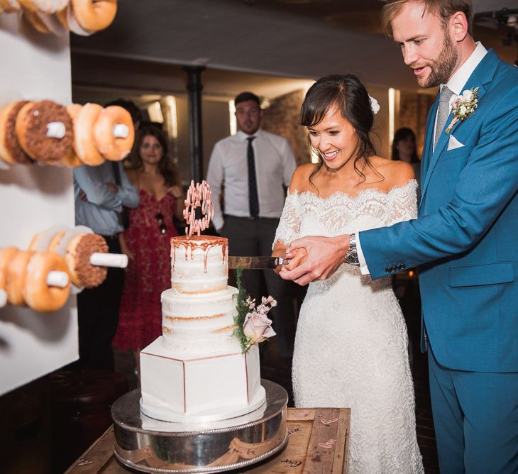 Bride in Lace Watters Dress with Bardot Neckline and Buttons Down Back | Bridal Up Do | Groom in Royal Blue Three-Piece Suit from Moss Bros. with Silver Tie and Pocket Square from T.M.Lewin | Three Tier Wedding Cake with Hexagon Bottom Layer and Semi-Naked Top Layers with Copper Drip Detail | Copper Calligraphy Cake Topper | Cutting of the Cake at The West Mill | Smoke Bombs and Chinese Paper Fans Backdrop with Bride in Bardot Dress | Twig &amp; Vine Photography