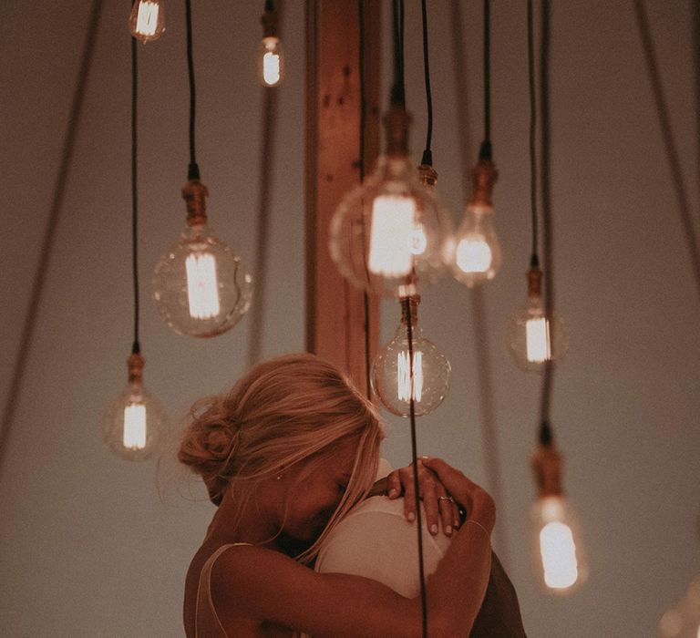 Bride in Rime Arodaky Wedding Dress with Lace Back and Groom in Morning Suit Embracing Under an Edison Light Bulb Installation