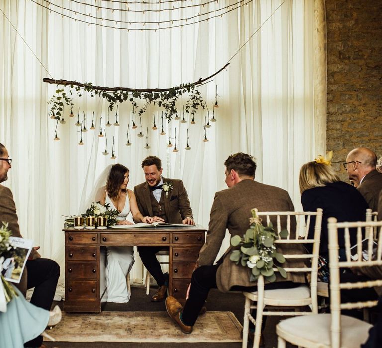 Bride and groom sign wedding register with hanging flowers behind